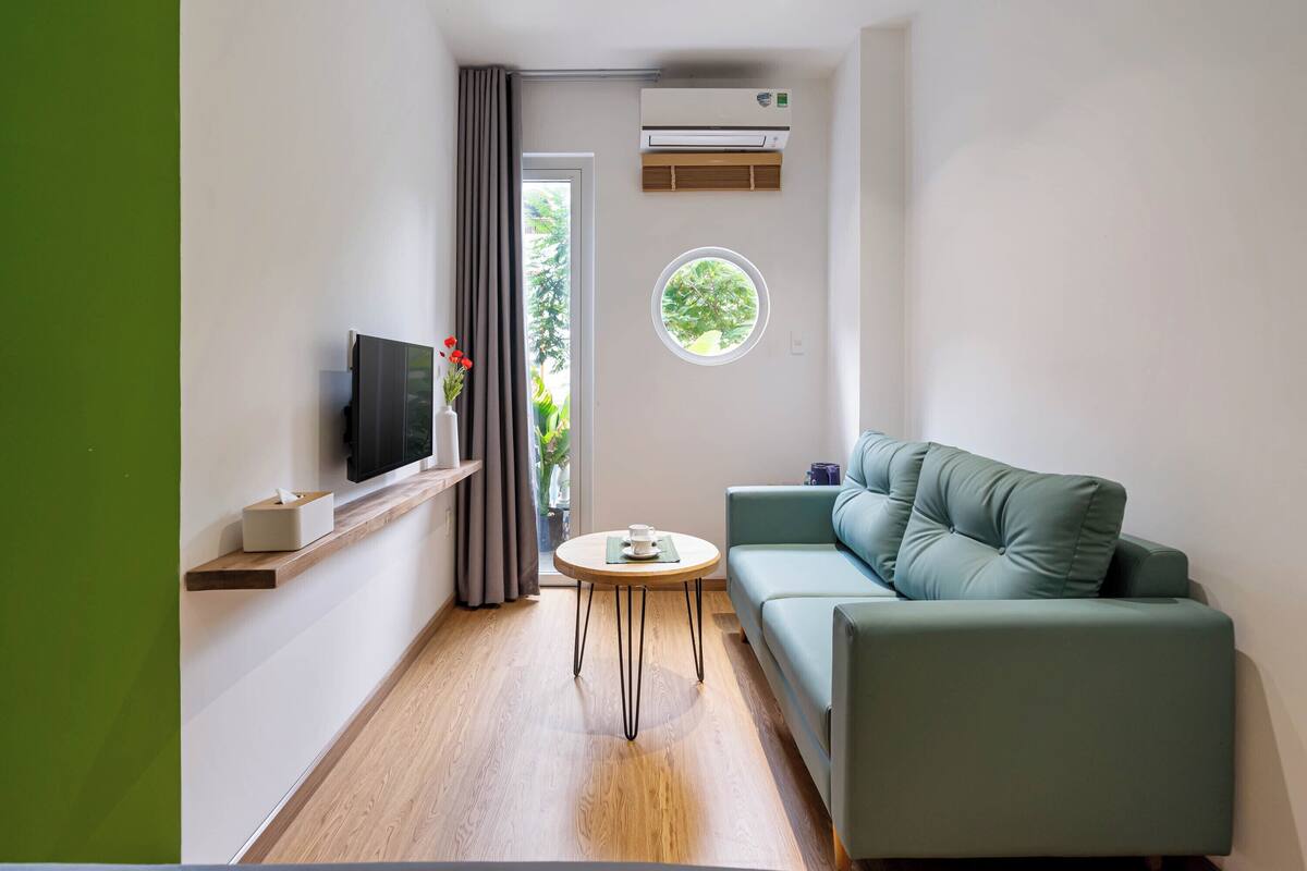 Een moderne woonkamer met een flatscreen televisie, een ronde tafel, een groene bank en een raam met uitzicht op groen.