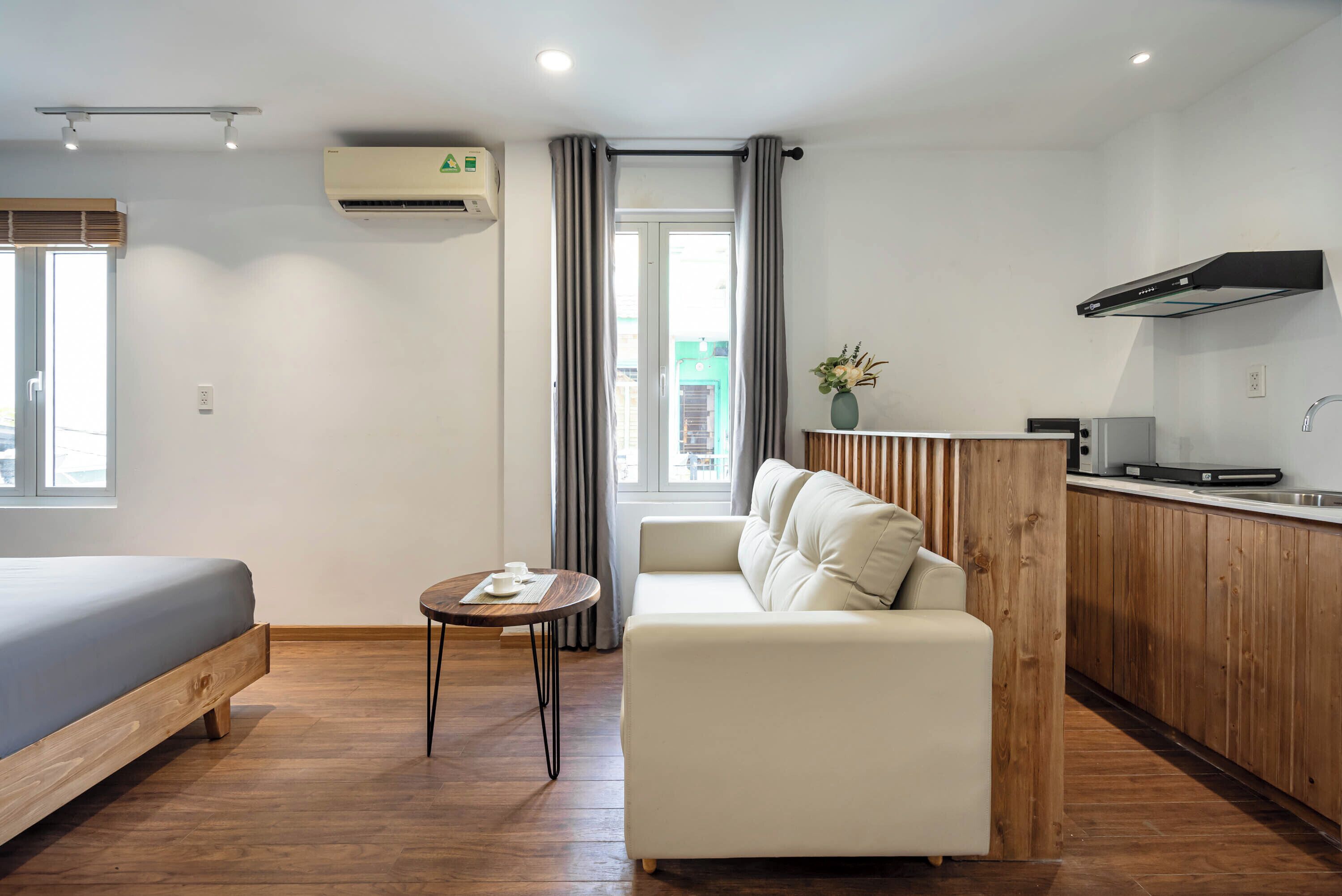 A modern hotel room with a bed, a sofa, a small round table, a kitchen area, and a window with curtains.