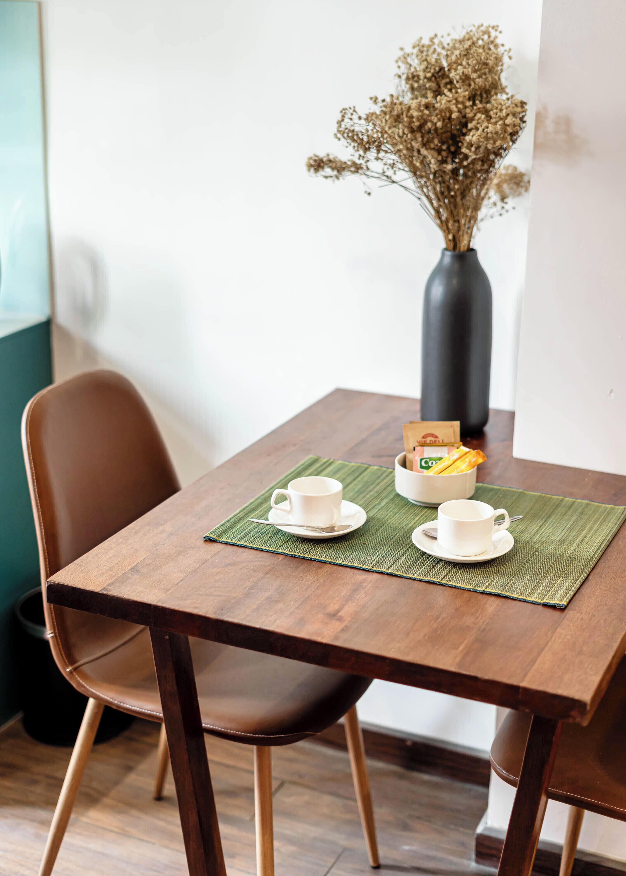A wooden table with two white cups and saucers, a green placemat, a vase with dried flowers, and a box of coffee.