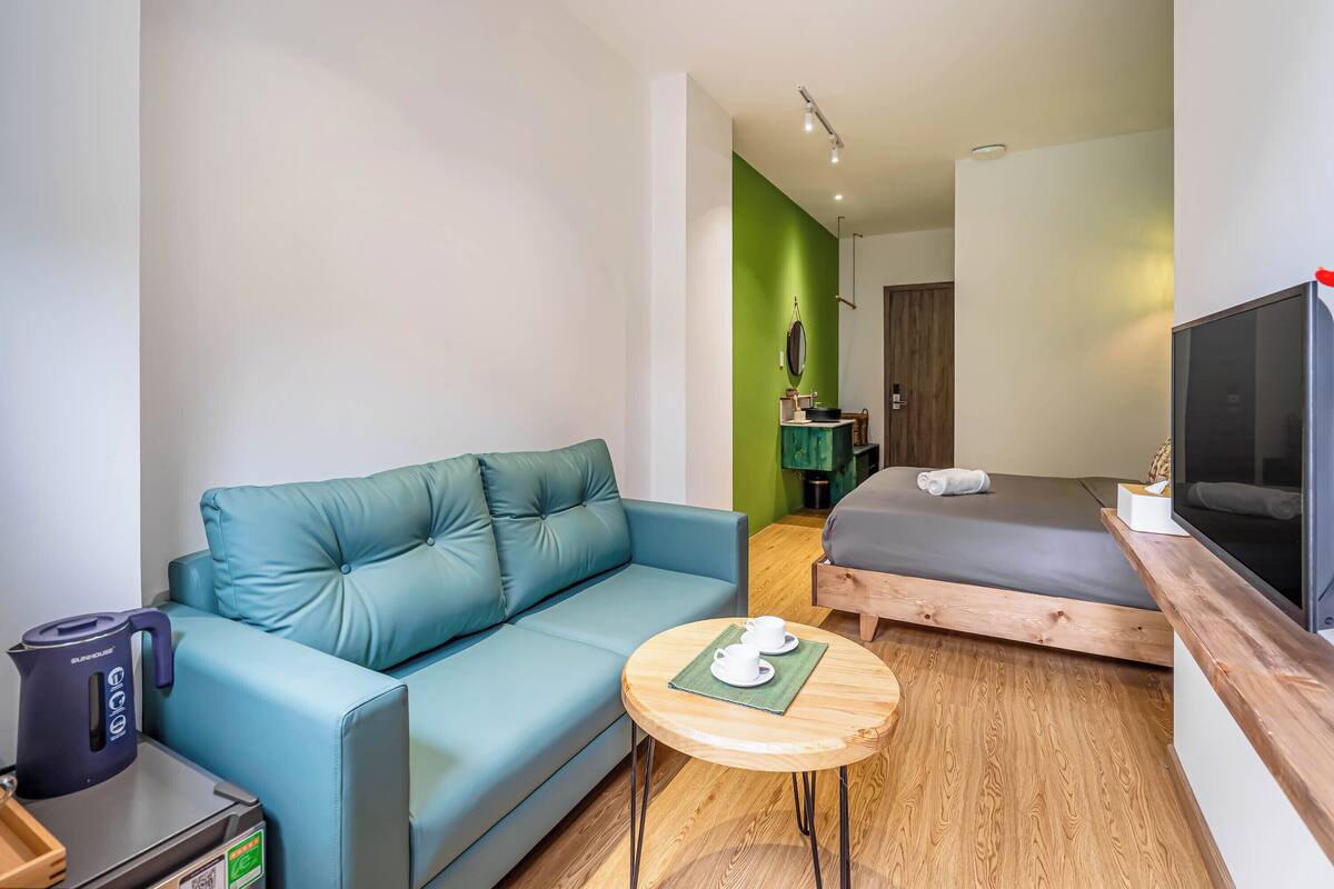 Een moderne hotelkamer met een blauwe bank, een klein rond houten tafeltje met een theepot en kopjes, een bed met een grijze matras, en een televisie op een houten dressoir.