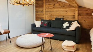 A cozy room with a dark grey sofa, a round white ottoman, a small red table, and a unique antler chandelier. - Spacious 4-Bedroom Apartment with Wi-Fi and Garage in Les Gets (Les Gets)