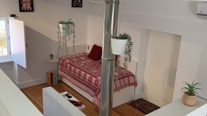A modern loft bedroom with a high ceiling, a large bed with a patterned red blanket, two hanging planters, and a white wardrobe. - House in granite stone with a century old in center Portugal. Open Space. (Tinalhas)