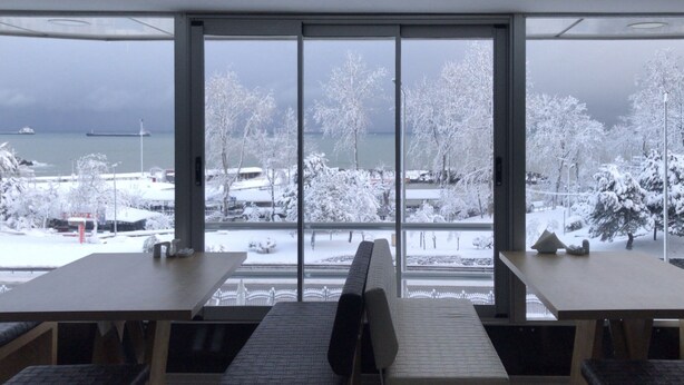 A room with a large window overlooking a snowy landscape with trees and a body of water. There are two tables and chairs in the room. - ALARGA PREMIER (Trabzon)