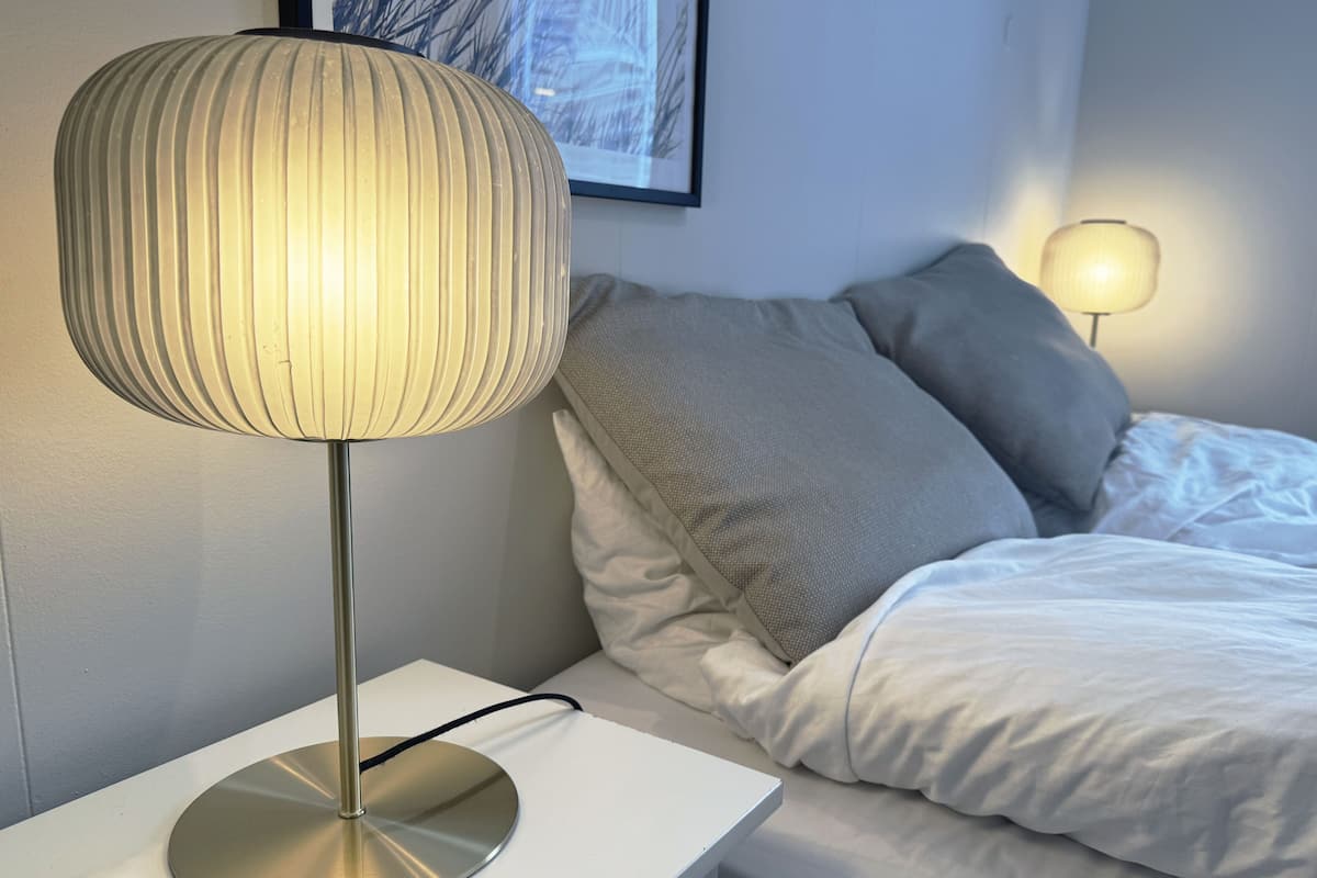 A modern bedroom with a bedside table, a lamp, a bed with pillows, and a framed picture on the wall.