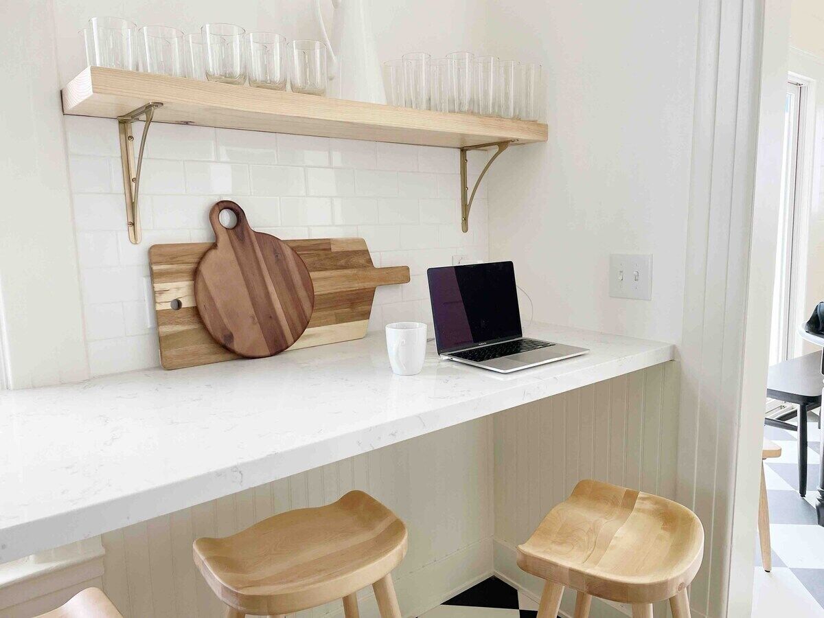 Une cuisine avec un plan de travail blanc, un ordinateur portable, une planche à découper en bois et deux tabourets en bois.