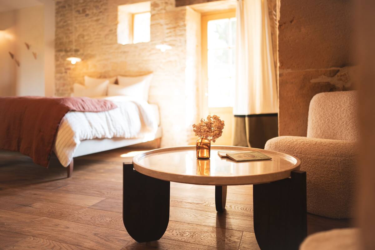 A cozy bedroom with a wooden floor, a round table with a vase, a bed with a red blanket, and a stone wall.