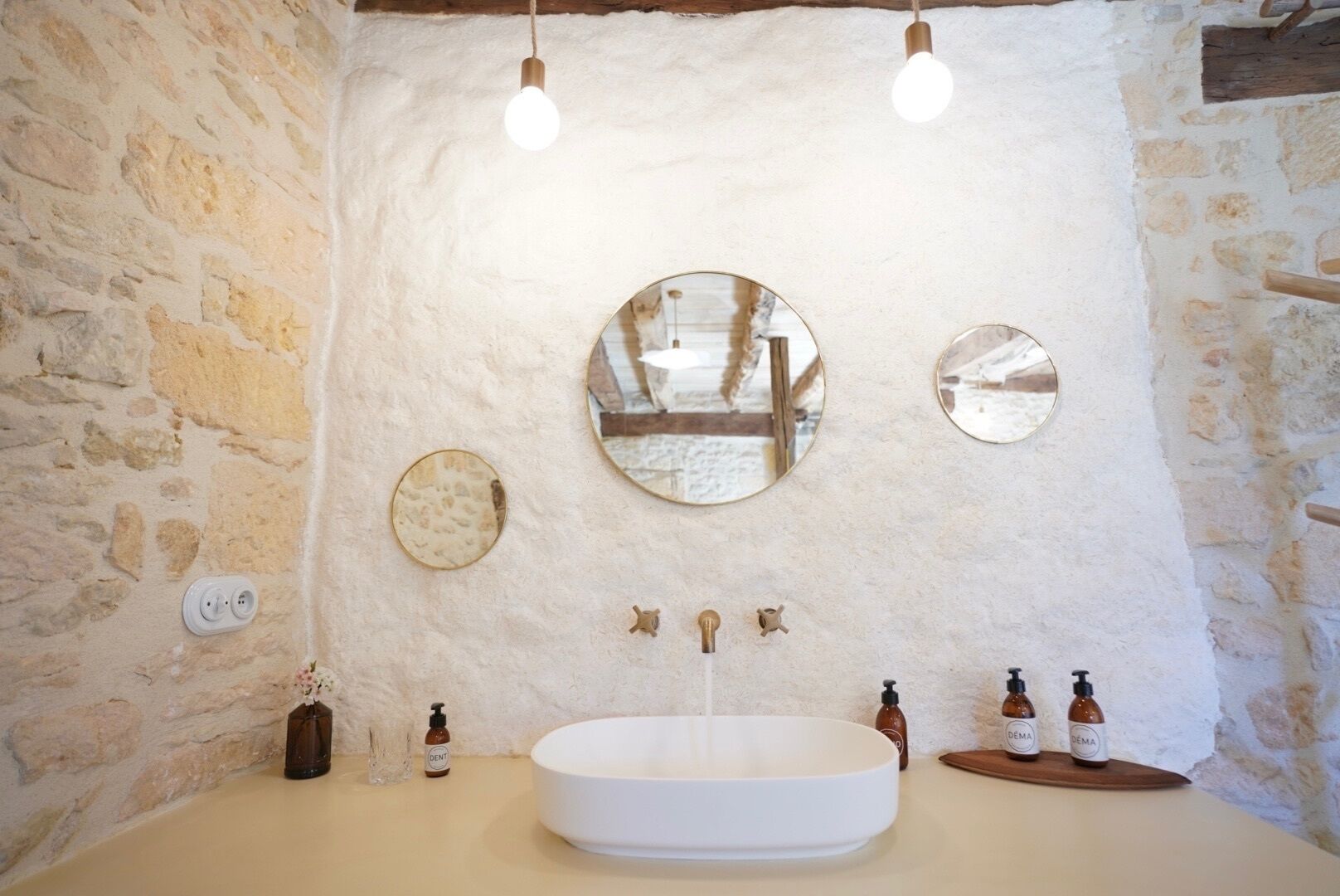 A bathroom with a stone wall, a round mirror, a sink, and two bottles of DEMA soap.