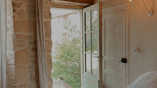 A bedroom with a stone wall, wooden beams, a bed with white bedding, a window with curtains, and a light fixture. - Maison Lou Piade (Sarlat-la-Canéda)