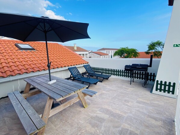 A rooftop patio with a wooden table, two lounge chairs, a barbecue grill, and a black umbrella.