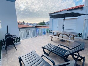 Eine Terrasse mit Grill, einem Holztisch und Liegestühlen.