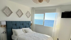 Ein Schlafzimmer mit Bett, Fenster mit Meerblick, Deckenventilator und einem an der Wand befestigten Fernseher.