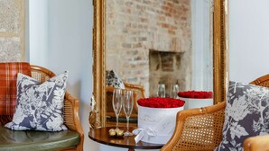 Una habitación acogedora con pared de ladrillo, una silla de madera con cojín floral, una mesa con un ramo de rosas rojas, dos copas de champán y un estuche decorativo.