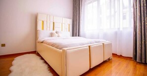 A bedroom with a large bed, a fluffy white rug, and a window with curtains. - Lux Suites Spring Garden Apartments (Nairobi)