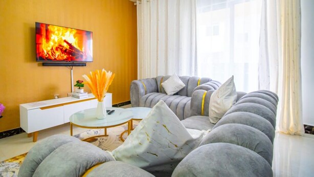 Ein Wohnzimmer mit einer Sofagarnitur, einem Couchtisch, einem an der Wand montierten Fernseher und einer Vase mit Blumen auf einem Beistelltisch.