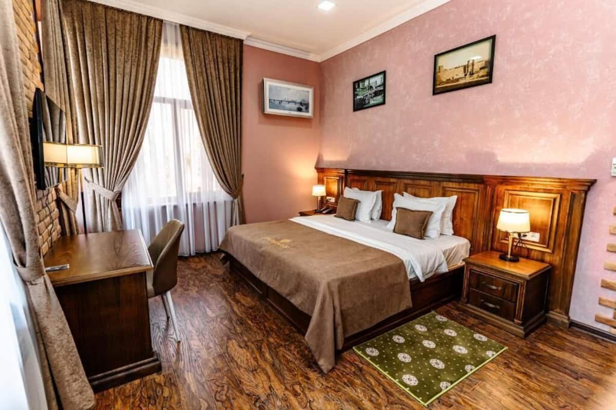 Ett hotellrum med en säng i trä, ett skrivbord, en stol och ett litet bord. Det finns två sänglampor och ett fönster med gardiner.