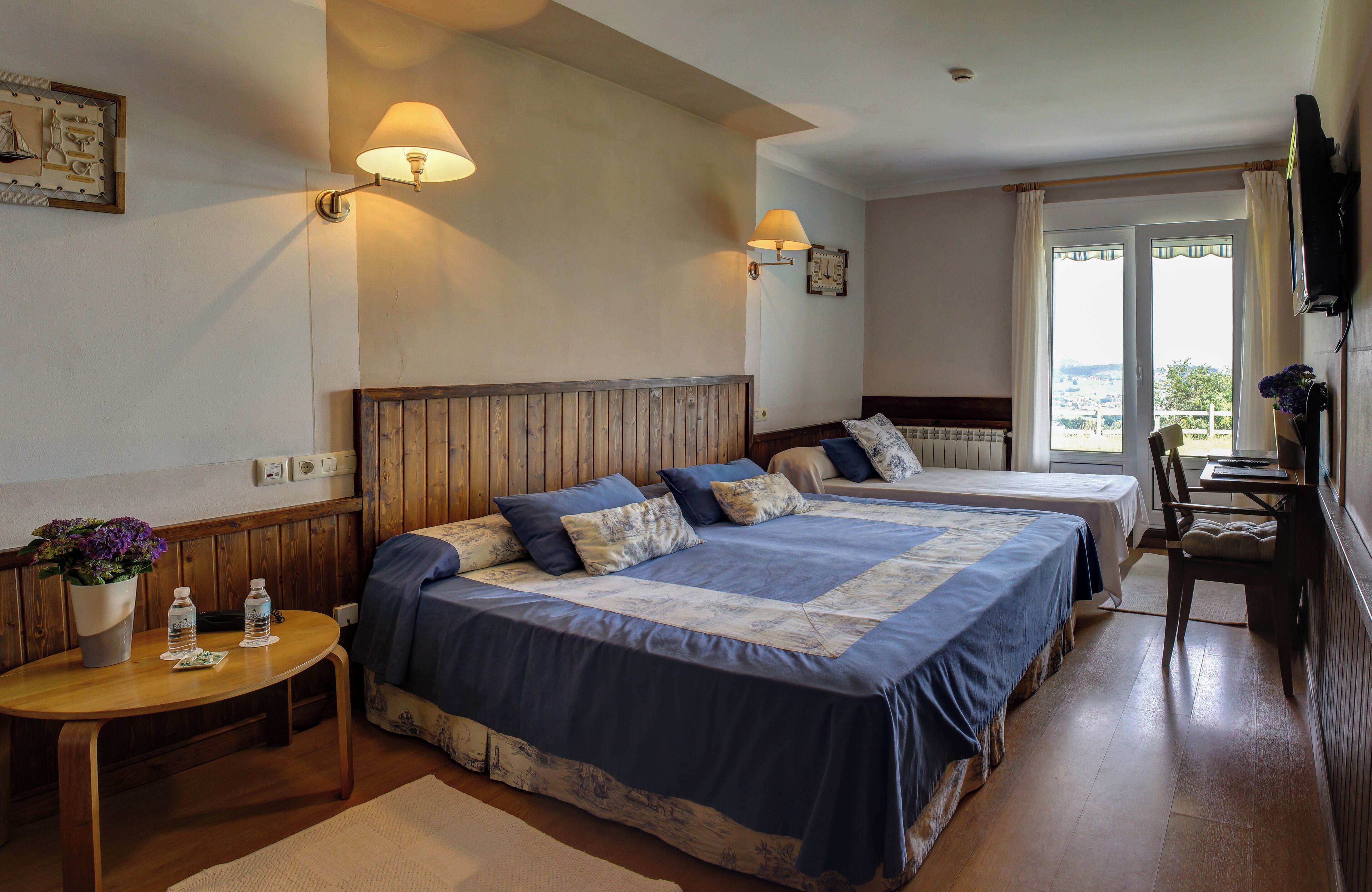 Une chambre d’hôtel avec deux lits, une tête de lit en bois, des lampes de chevet, une petite table avec une plante et une vue par la fenêtre.