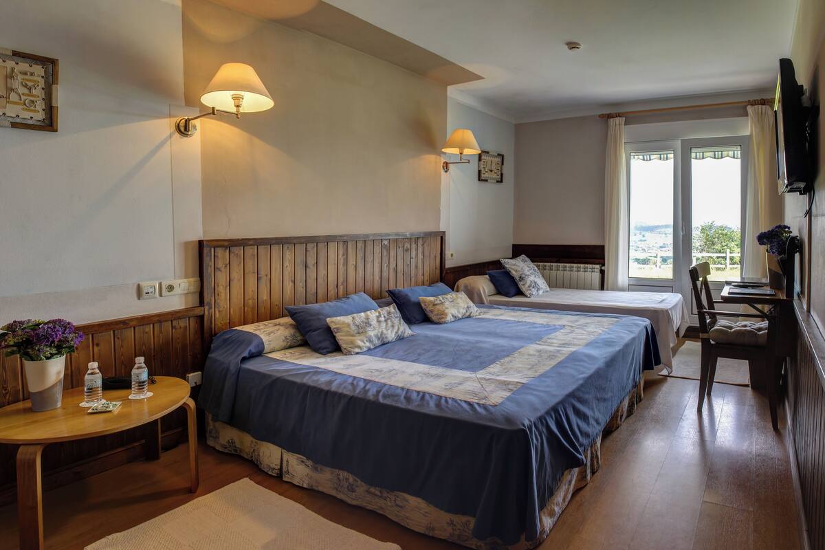 Een hotelkamer met twee bedden, een houten hoofdbord, nachtlampjes, een kleintje tafeltje met een plant en uitzicht door een raam.