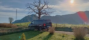 Un vehículo negro estacionado sobre un sendero cubierto de hierba, con montañas al fondo.