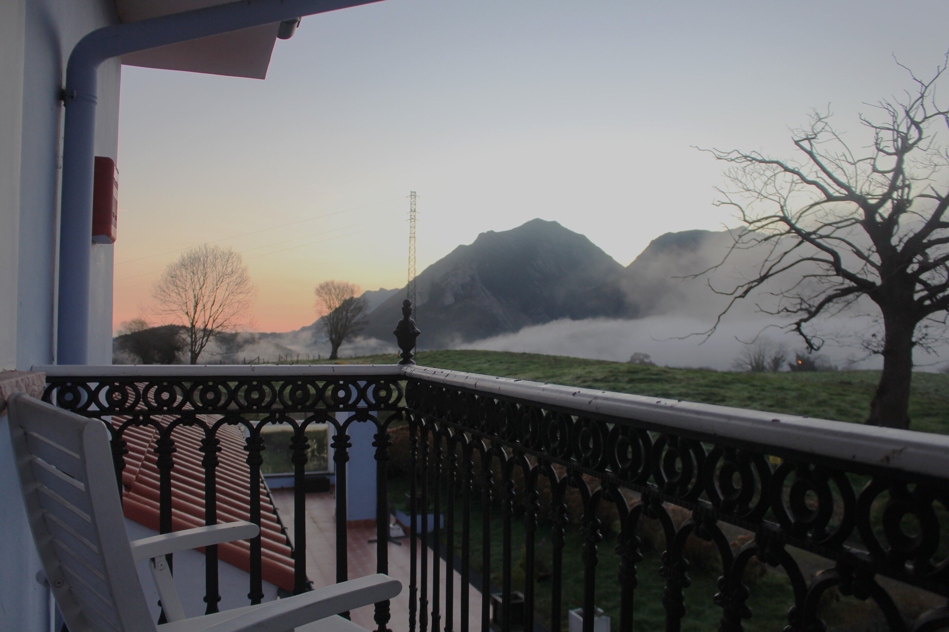 A balcony with a metal railing, a bench, and a view of mountains and a tree in the background.