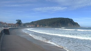 A beach with a curved shoreline, a stone wall, and a forested hill in the background. - Hotel Mirador del Sella (Ribadesella)