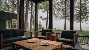 A modern outdoor seating area with a wooden table and sofas, overlooking a grassy landscape and trees. - Clay Banks Lakehouse · Door County Lake House 50 min from Lambeau Field (Algoma)