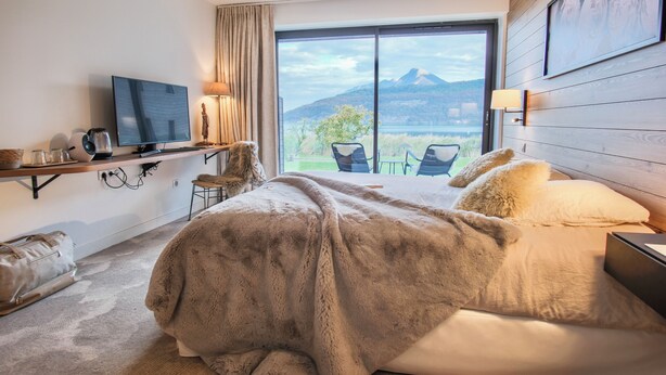 Quarto de hotel com cama, televisão, escrivaninha com luminária e vista para montanhas através de uma janela grande.