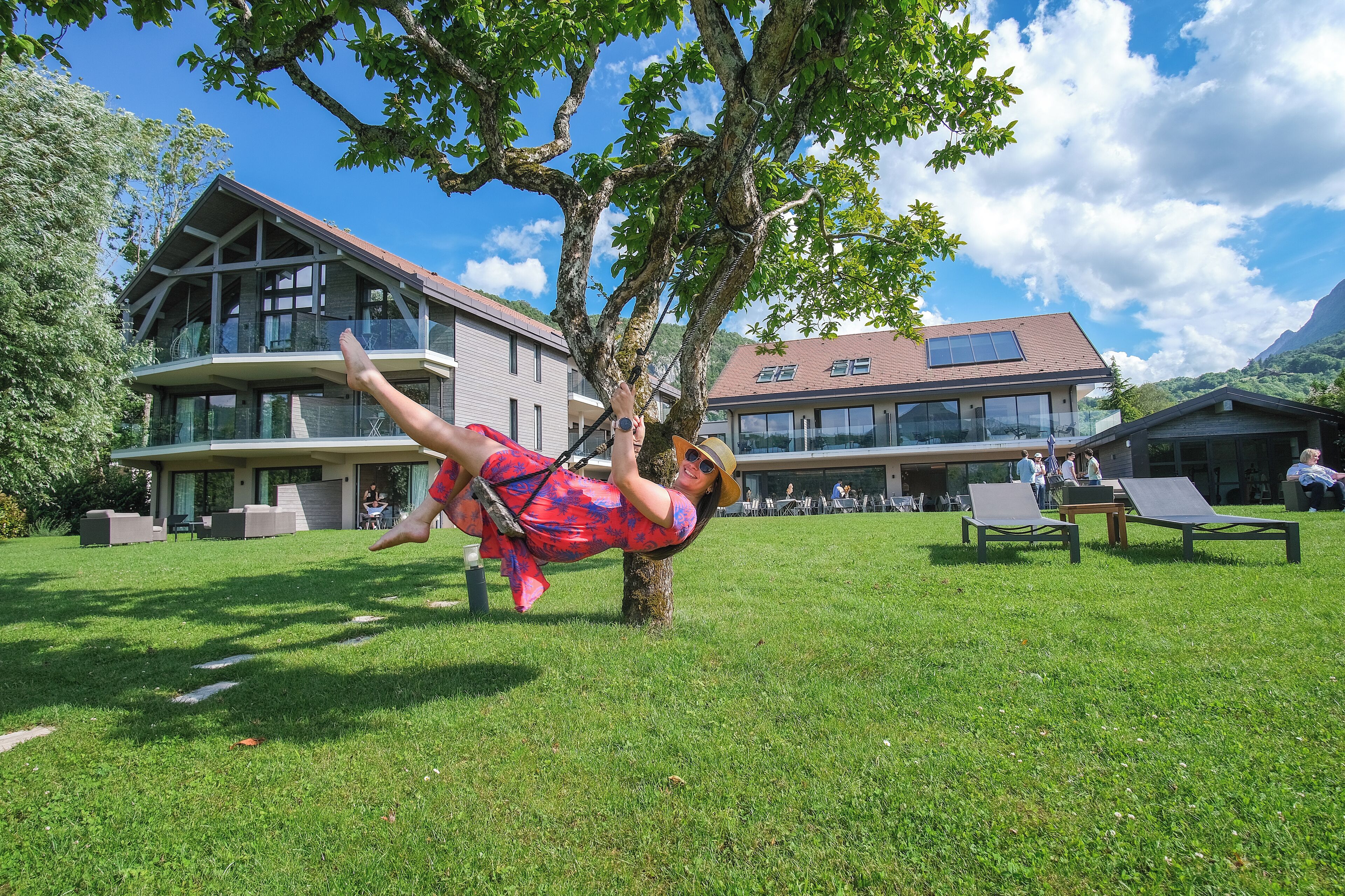 A person relaxing on a swing under a tree in a grassy area with a building in the background.