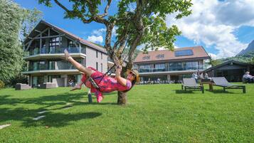 A person relaxing on a swing under a tree in a grassy area with a building in the background.