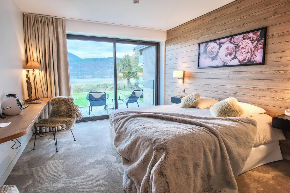 A hotel room with a large bed, a desk with a chair, a window with a view of the outdoors, and a wooden wall with a framed picture.
