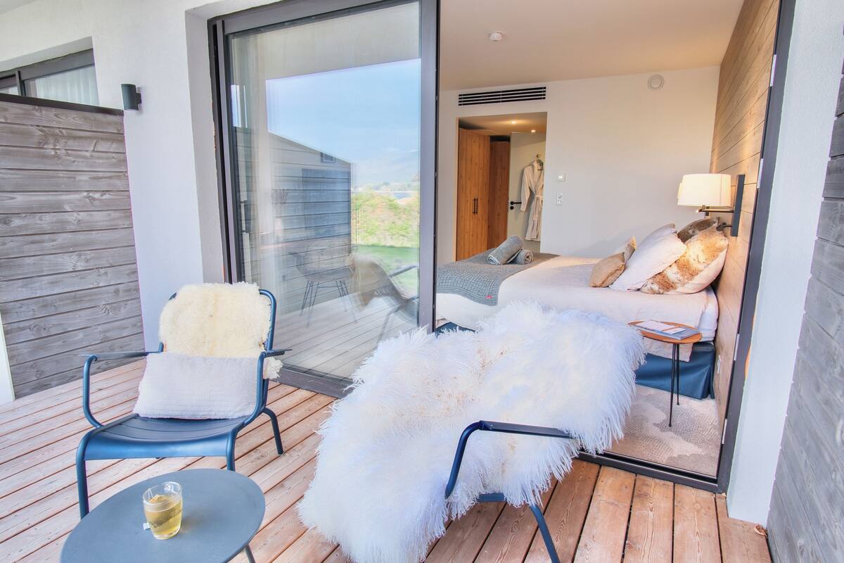 A modern bedroom with a large sliding glass door leading to a balcony, a bed with fluffy white bedding, a small table with a drink, and a chair with a fluffy cushion.