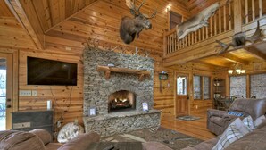 L’intérieur d’une cabane en rondins, avec une cheminée en pierre, des meubles en cuir, des têtes d’animaux empaillées et un téléviseur.