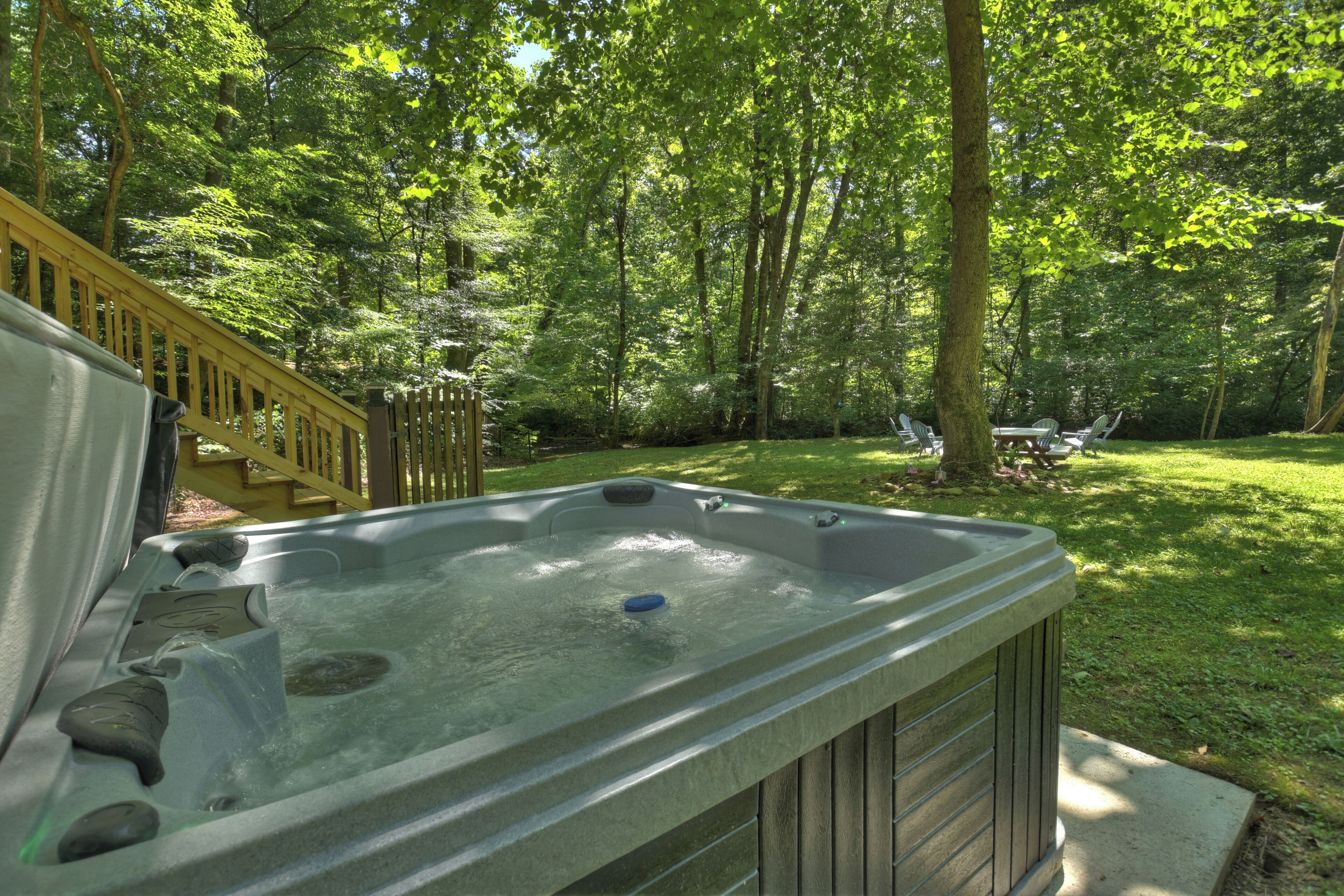 Un jacuzzi en un patio trasero arbolado con una terraza de madera y una mesa de picnic.