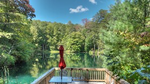 A wooden deck with a table and chairs overlooking a lake, surrounded by dense forest. - Amazing Mountain Views! Fenced Yard, Hot Tub, Game Room with Arcade Game & Pool Table, Firepit, Fishing Pond - View Point (Blue Ridge)