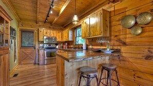 A wooden cabin kitchen with a central island, bar stools, and a hanging light fixture. - Amazing Mountain Views! Fenced Yard, Hot Tub, Game Room with Arcade Game & Pool Table, Firepit, Fishing Pond - View Point (Blue Ridge)