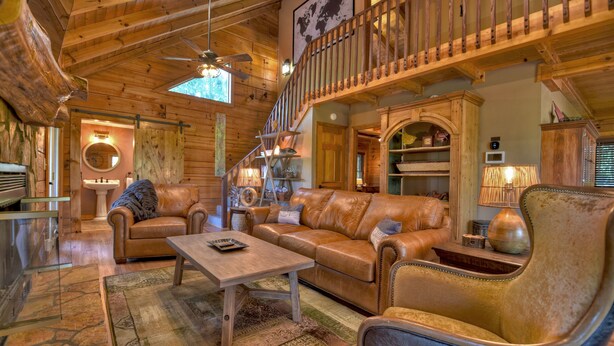 A cozy log cabin interior with a wooden staircase, leather sofas, a dining table, and a large window. - Amazing Mountain Views! Fenced Yard, Hot Tub, Game Room with Arcade Game & Pool Table, Firepit, Fishing Pond - View Point (Blue Ridge)