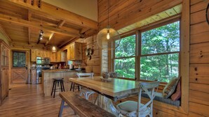 A rustic cabin interior with wooden walls, a large wooden dining table, and a view of the forest through a large window. - Amazing Mountain Views! Fenced Yard, Hot Tub, Game Room with Arcade Game & Pool Table, Firepit, Fishing Pond - View Point (Blue Ridge)