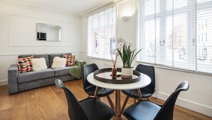 A modern living room with a grey sofa, black chairs, a round table, and large windows with blinds. - Charming Flat in Covent Garden (Greater London)
