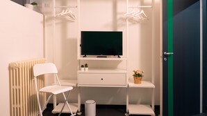 Een kleine kamer met een wit bureau, een televisie, een stoel, een radiator en een deur met een groene streep.