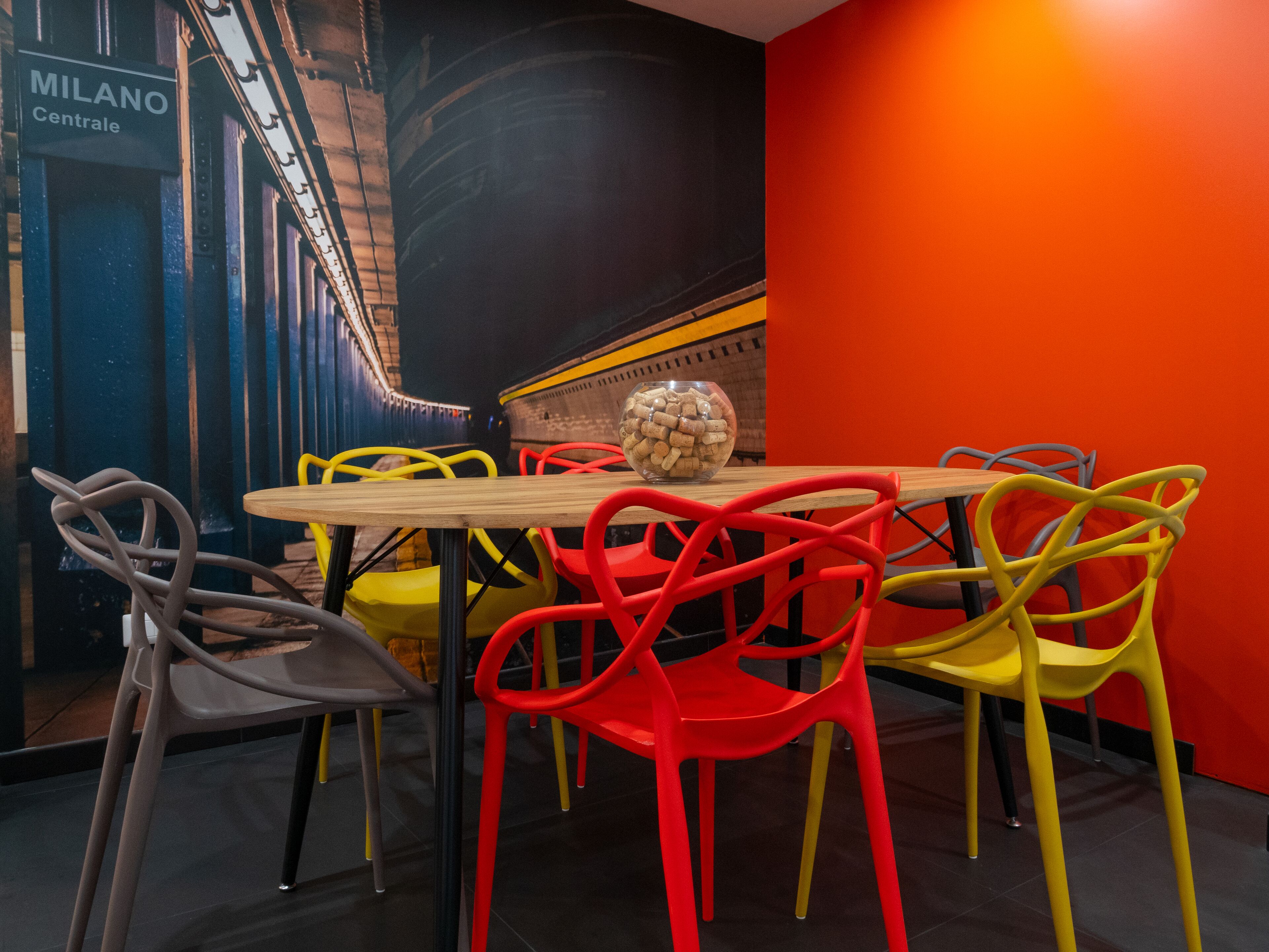 Un coin repas avec une table en bois et des chaises colorées, un bol de collations et un panneau indiquant « MILANO Centrale ».