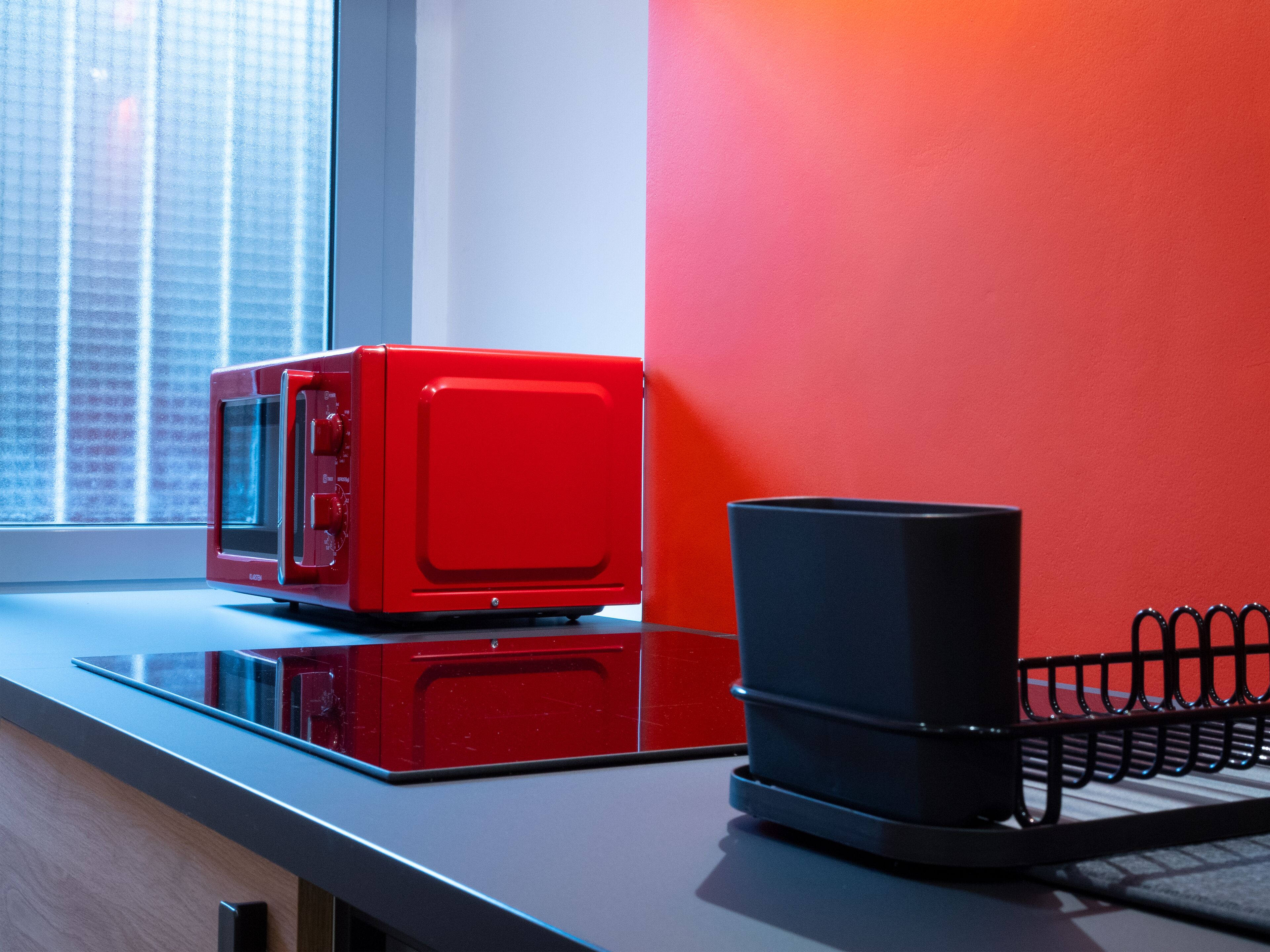 Un forno a microonde rosso su un piano di lavoro in cucina, accanto a una tazza da caffè nera e una scaffalatura in metallo nero.