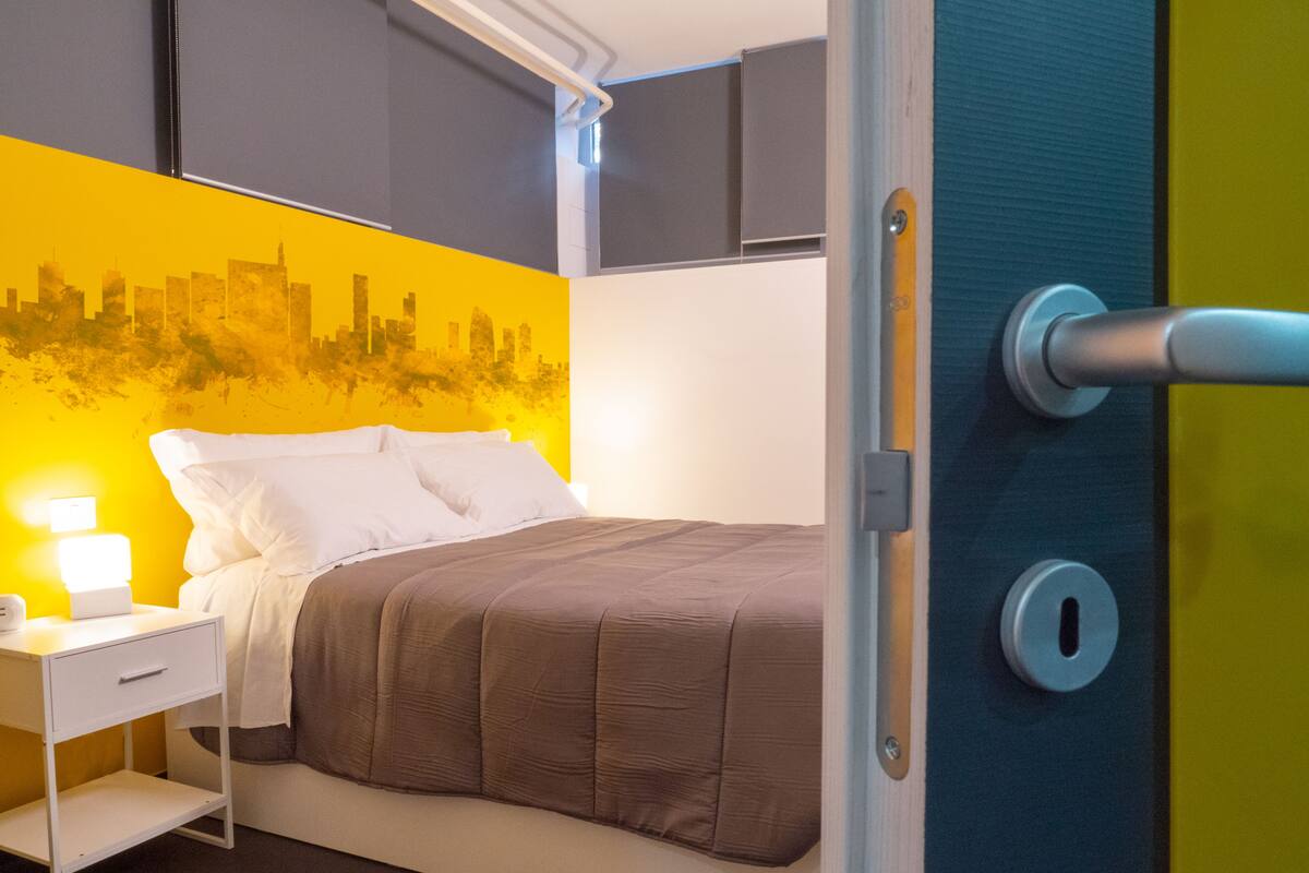 Una cama bien hecha con una cubierta marrón, una mesita de noche blanca con una lámpara y un mural de paisaje urbano en la pared.
