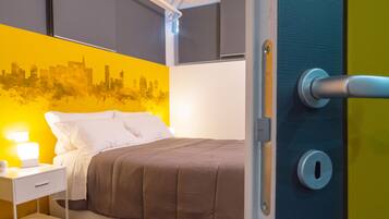 A neatly made bed with a brown cover, a white bedside table with a lamp, and a cityscape mural on the wall.