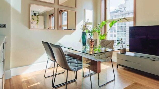 Un salon moderne avec une table à manger en verre, deux chaises, un téléviseur à écran plat et une grande fenêtre donnant sur la ville.