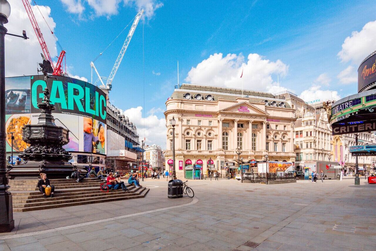 A public square with a large building featuring classical architecture, a prominent advertisement for 'GARLIC', and a statue.