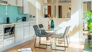 Une cuisine moderne avec des meubles blancs, un pare-feu turquoise et un coin repas comprenant une table en verre et des chaises en métal.