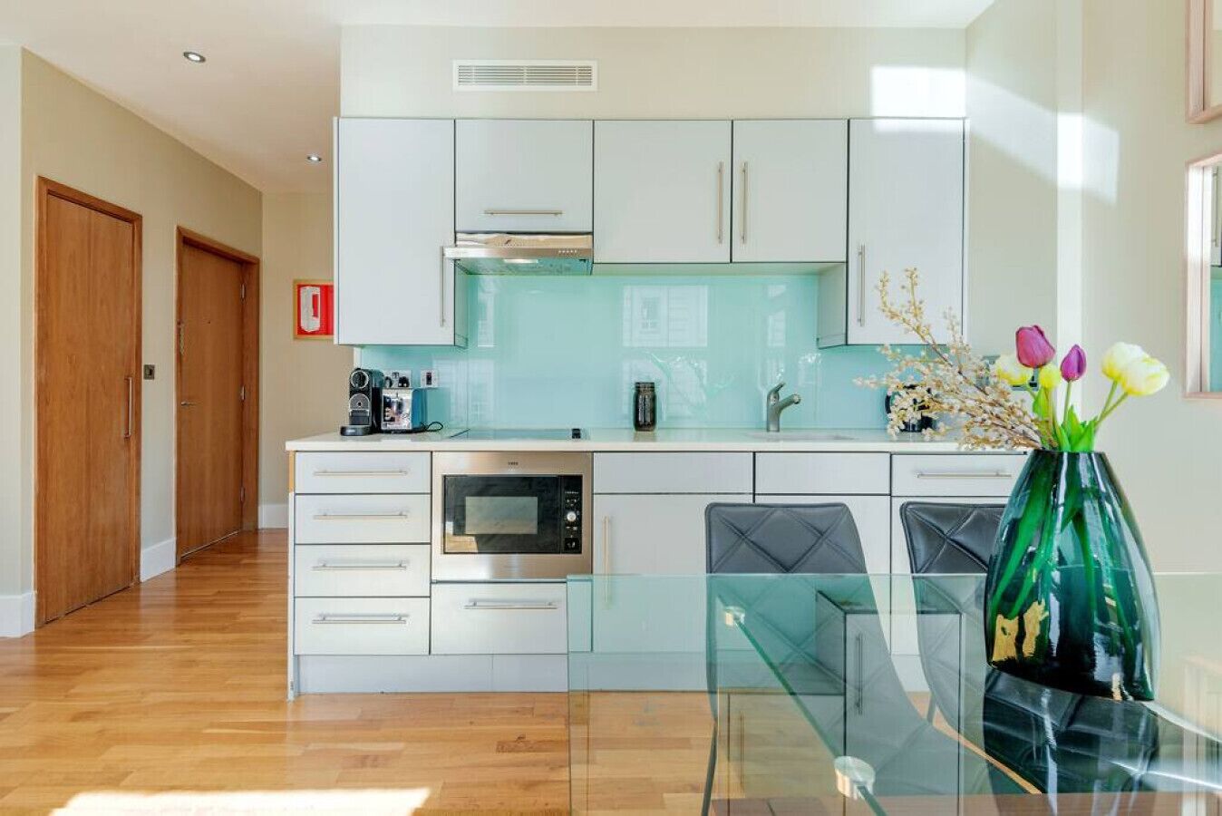 A modern kitchen with white cabinets, a glass countertop, a built-in oven, and a coffee machine.