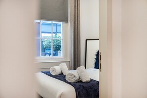 A neatly made bed with rolled white towels, a dark blue throw blanket, and a window with curtains.