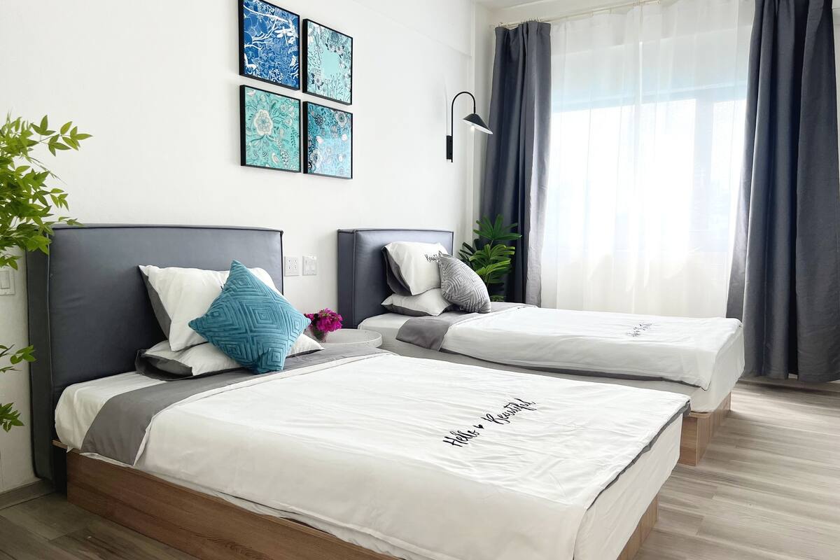 A modern hotel room with two single beds, a large window with curtains, and wall art.
