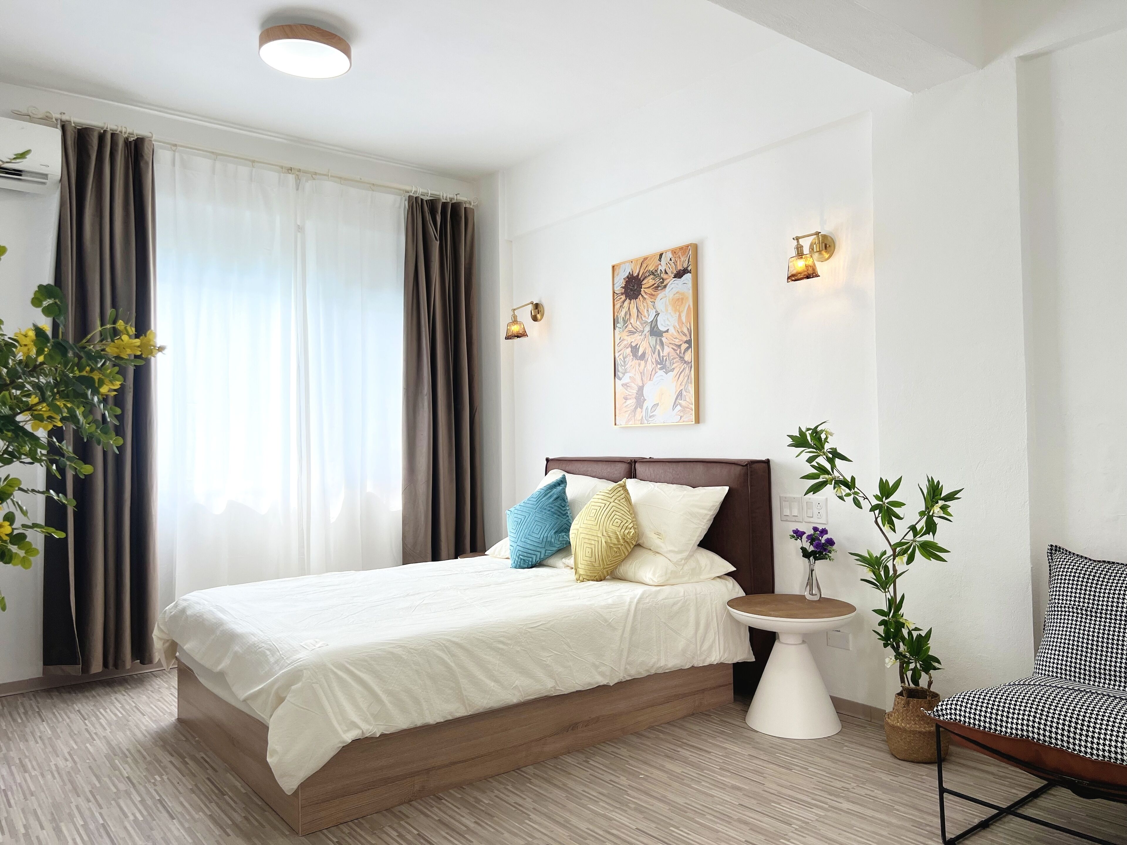 Quarto de hotel com cama, mesa de cabeceira, cadeira, planta em vaso e um quadro na parede.