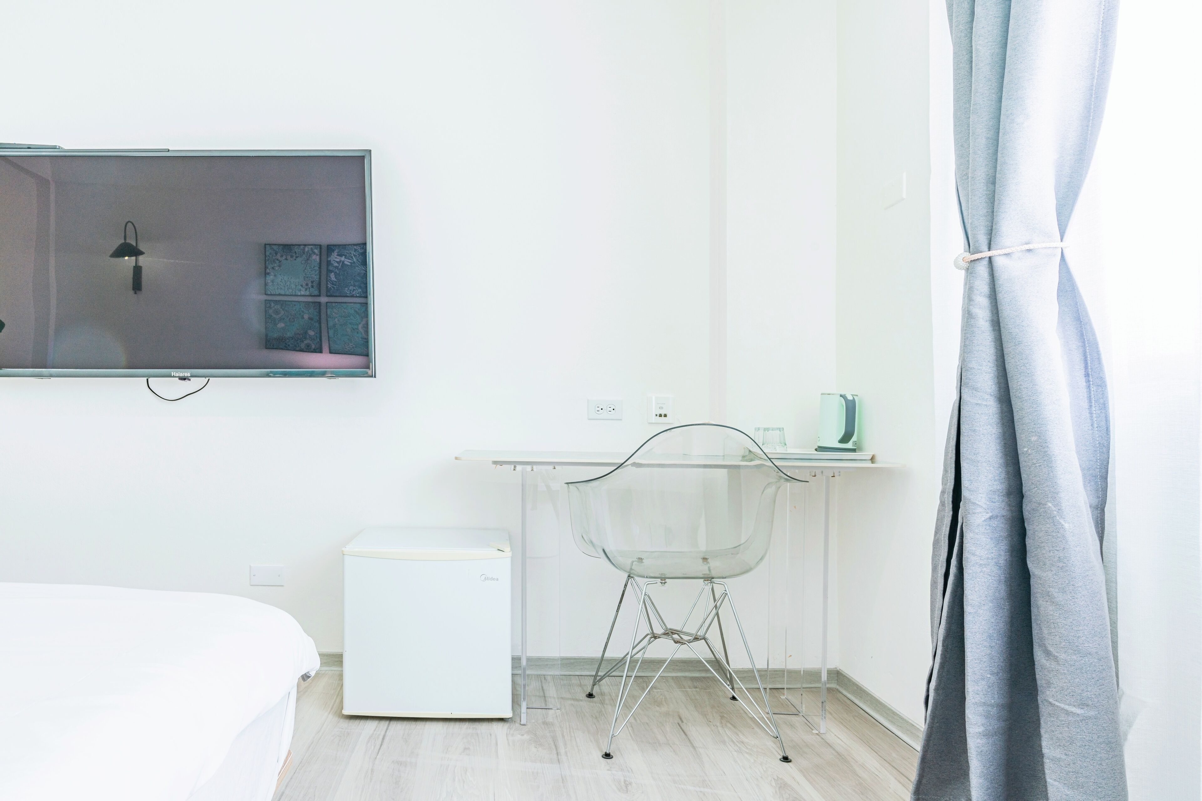 Une chambre d’hôtel minimaliste comprenant un lit, un petit réfrigérateur blanc, une table en verre avec une chaise, un téléviseur à écran plat et un rideau.